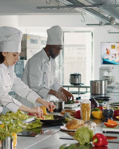 Team of cooks slicing vegetables on cutting board for meal preparation in restaurant kitchen. Man and woman cooking gourmet food dish with organic ingredients, working on culinary recipe.